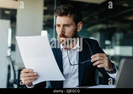 Focused businessman reading negative bills, making business accounting reports, sitting in office interior, copy space Stock Photo