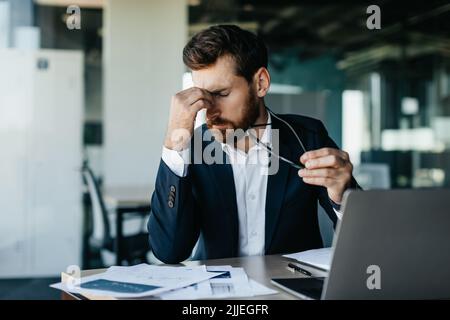 Exhausted businessman sitting in front of laptop at modern office, holding glasses and rubbing nose bridge Stock Photo