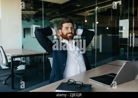 Relaxed happy businessman sitting at workplace in office, leaning back on chair and smiling, free space Stock Photo