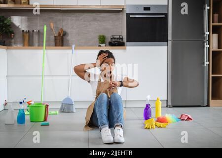 Shocked young black housewife with open mouth sit on floor with cleaning supplies, look at clock Stock Photo