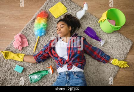 Tired millennial african american woman housewife lying down on floor with cleaning supplies in room interior Stock Photo
