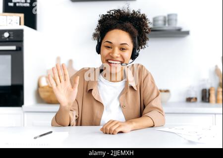 Positive confident curly-haired young african american woman with headset, call center worker, consultant, talks via video call, female mentor recording webinar, lecture, gesturing with hands, smiling Stock Photo