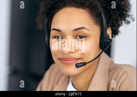 Close-up photo of pretty, confident curly-haired young african american woman with headset, call center worker or consultant, wearing stylish shirt, looks directly at the camera, smiling Stock Photo