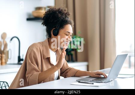 Positive successful african american curly-haired young woman, in casual clothes, working or studying from home, using laptop and headset, talking on video conference, smiling friendly Stock Photo