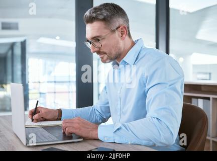 You have to stay focused on what you want. a mature businessman writing notes while working on a laptop in an office. Stock Photo