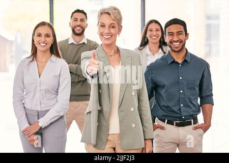 You can trust us to get the job done. an attractive mature businesswoman standing in the office with her colleagues and showing a thumbs up. Stock Photo