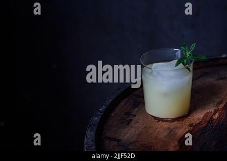 glass of ginger beer Moscow mule with large ice and lemon verbena herb garnish on wood barrel dark background