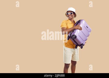 Happy afro american male tourist or traveler holding suitcase on beige background. Stock Photo
