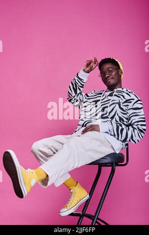 Portrait of African teenage boy in stylish clothes sitting on chair and posing at camera against pink background Stock Photo