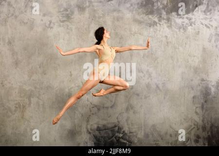 Full body side view of female ballet dancer in bodysuit leaping up with spread arms against gray wall in studio Stock Photo