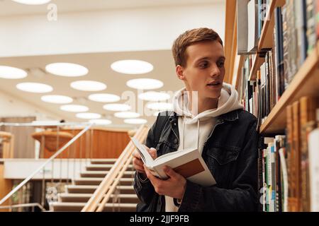 Young student with book looking at bookshelf. Teenager in library reading a book. Stock Photo