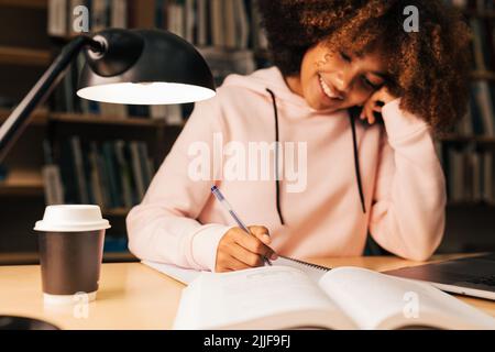 Cheerful student writing on notebook while sitting at desk in library Stock Photo