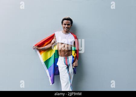 Happy guy with rainbow flag standing outdoors at grey wall and looking at camera wearing stylish clothes Stock Photo