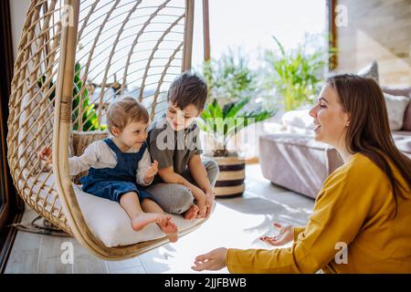 Young cheerful mother playing with her little children and having fun when swinging them on hanging chair in conservatory at home. Stock Photo