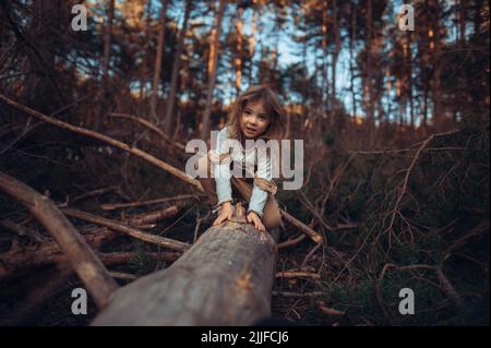 Autumn portrait of happy little girl balancing on tree trunk outdoors in forest, looking at camera. Stock Photo