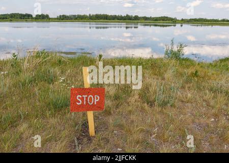 On the shore of the lake is a stick stuck with a red warning Sign Stop. Walking near the water and swimming is prohibited. Stock Photo