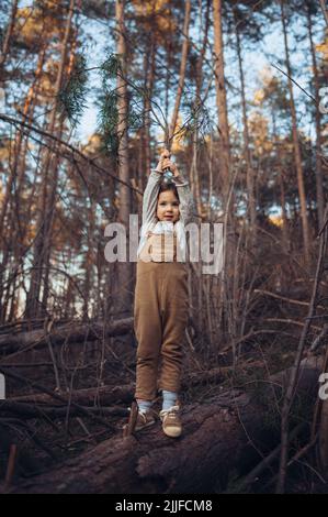 Autumn portrait of happy little girl balancing on tree trunk with raised arms outdoors in forest. Stock Photo