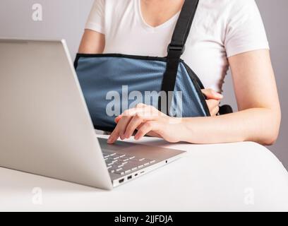 Woman with painful arm in sling using laptop for work, study. Female suffering from shoulder, clavicle ache, sitting at table with computer. High quality photo Stock Photo