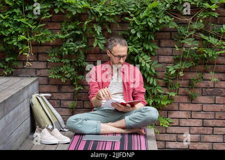 Young relaxed student guy sitting in lotus pose reading book outdoors, enjoying studying outside Stock Photo