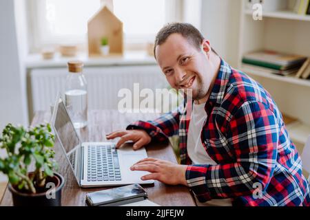 Young man with Down syndrome sitting at desk in office and using laptop, looking at camera and smiling. Stock Photo