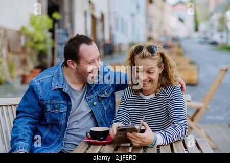 Portrait of happy young man with Down syndrome with his mother sitting at cafe outdoors and talking. Stock Photo