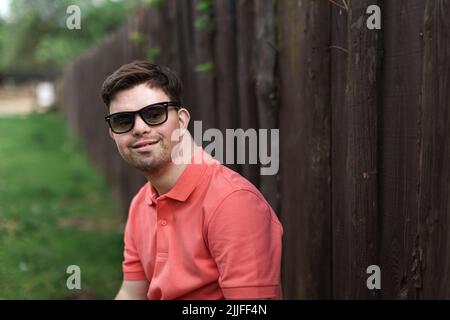 Portrait of happy young man with down syndrome standing outdoors in park and wearing sunglasses Stock Photo