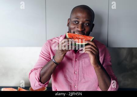 Multiracial man eating watermelon in his kitchen during hot sunny days. Stock Photo