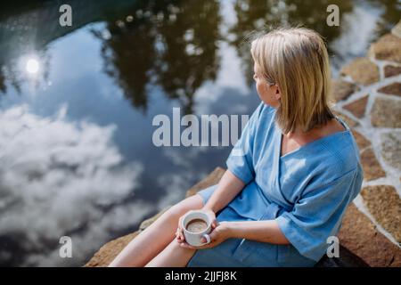 Happy young woman sitting by pond near cottege and enjoying cup of morning coffee on summer vacation in mountains. Stock Photo