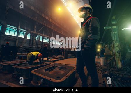Engineer in helmet, face mask and uniform controls workers in workshop of Steel Mill. Heavy industry Foundry. Stock Photo