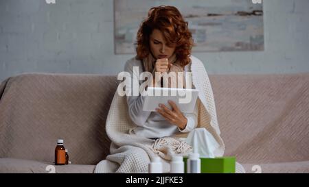 redhead sick woman using digital tablet and coughing near medication while sitting on couch Stock Photo