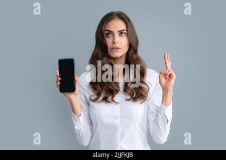 Happy young woman showing empty smartphone screen, crossed fingers for good luck. Portrait of her she nice attractive lovely focused girl holding Stock Photo