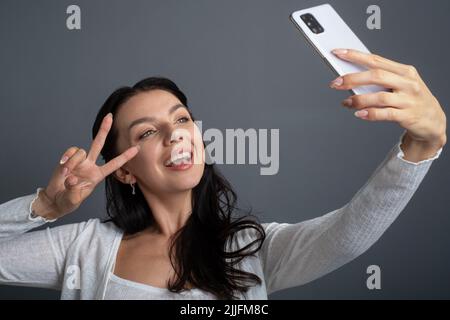 Taking selfie photos on cellphone. Young woman laughing and showing a victory peace gesture sign to a  front mobile phone camera. Stock Photo