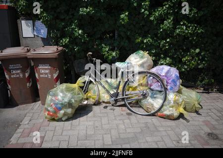 Parma, Italy - July, 2022: Old bicycle with basket on handlebar and broken wheel among garbage bags with plastic near brown trash cans. Discarded bike Stock Photo