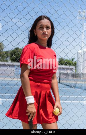 Young Woman with racket and ball standing near tennis court fence during break in training. Stock Photo