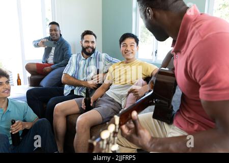 Multiracial happy male friends with beer bottles and cellphone looking at man playing guitar at home Stock Photo