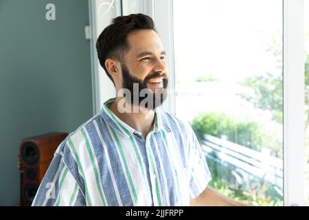 Smiling bearded caucasian young man looking through window while standing at home, copy space Stock Photo