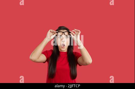 Surprised and shocked caucasian girl with eyeglasses, with eyes widened and mouth opened, holding glasses with hands on forehead, over red background