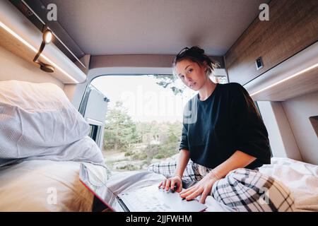Young female digital nomad in pajama lying on bed and browsing data on netbook while working remotely during trip in RV Stock Photo