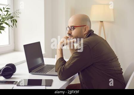 Pensive man sitting at his working desk with laptop, tablet, mobile phone and headphones Stock Photo