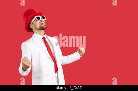 Happy emotional young man in suit laughs wildly and rejoices celebrating victory on red background. Stock Photo
