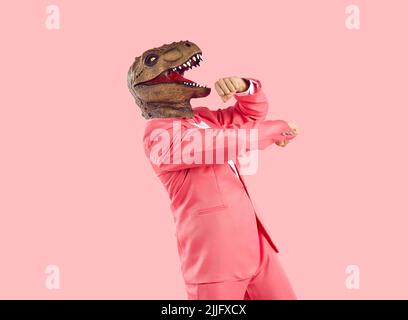 Cheerful funny and humorous man in dinosaur rubber mask dances isolated on pastel pink background. Stock Photo