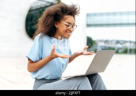 Busy positive young african american woman with glasses, mixed race female sitting outdoors, emotionally talking on video conference using laptop, gesturing with hands, looking at screen, smiling Stock Photo