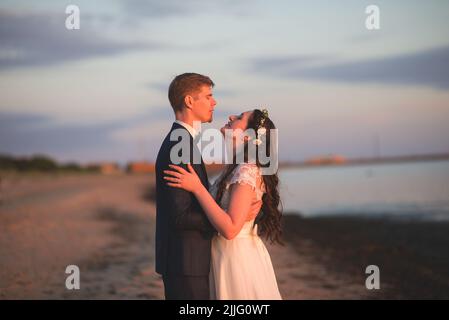 A young romantic bride and groom posing embracing on a beach at sunset Stock Photo