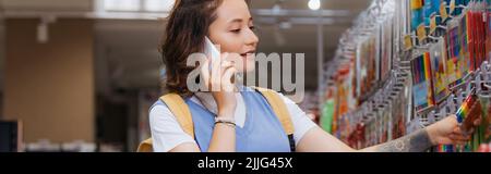 brunette woman choosing felt pens while talking on smartphone in stationery store, banner Stock Photo