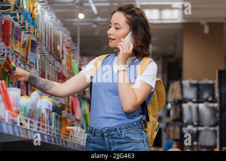 tattooed woman choosing felt pens while calling on cellphone in stationery shop Stock Photo