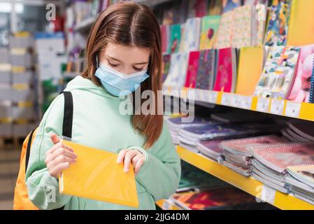 child in medical mask holding notebook while choosing school supplies in shop Stock Photo