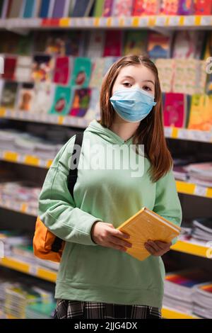 girl in medical mask holding notebooks near blurred rack with school supplies Stock Photo