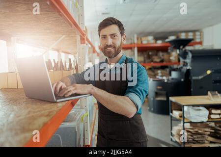 Smiling warehouse worker in uniform standing on coffee small factory with laptop Stock Photo