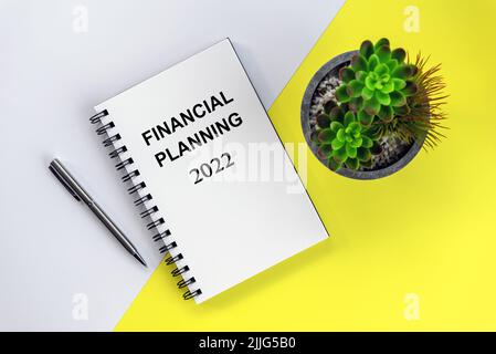 Financial planning 2022 in business flat lay concept with spiral notebook, pen and plant on white and yellow table background. Stock Photo