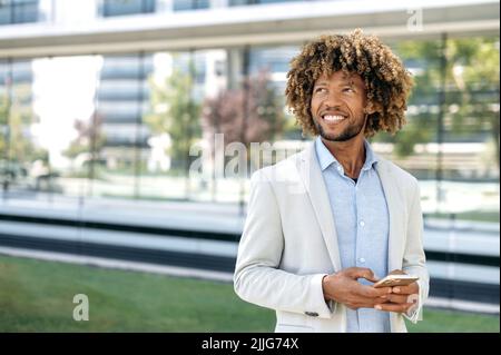 Successful positive brazilian or african american curly haired man, wearing stylish formal clothes, stands outdoors near business center, uses smartphone, typing, looks away, thinking, smiling happily Stock Photo
